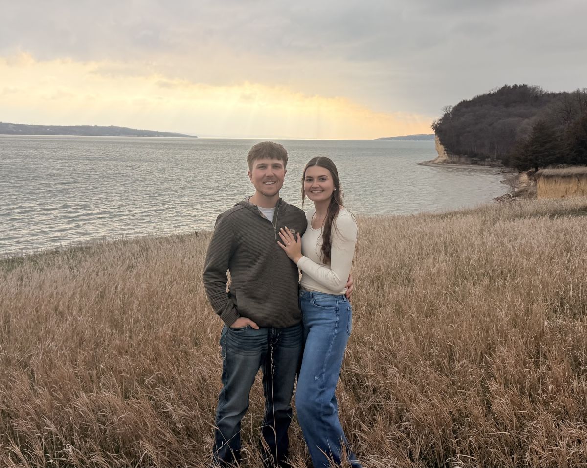 Couple smiling after a lakeside proposal at sunset, woman showing her engagement ring on a grassy bluff