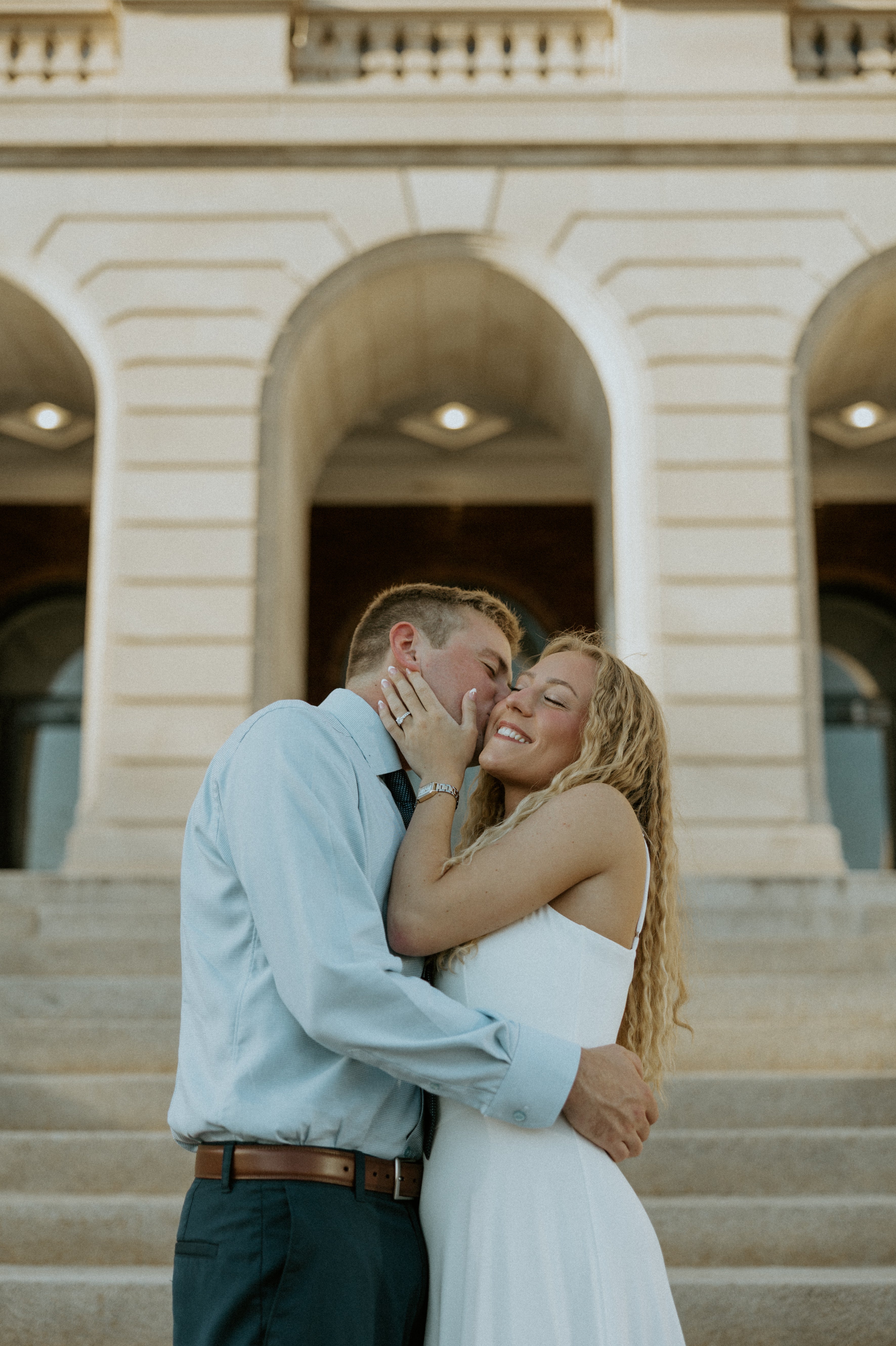 Couple embracing on stone steps, man kissing woman’s cheek as she smiles and shows engagement ring, arched building behind