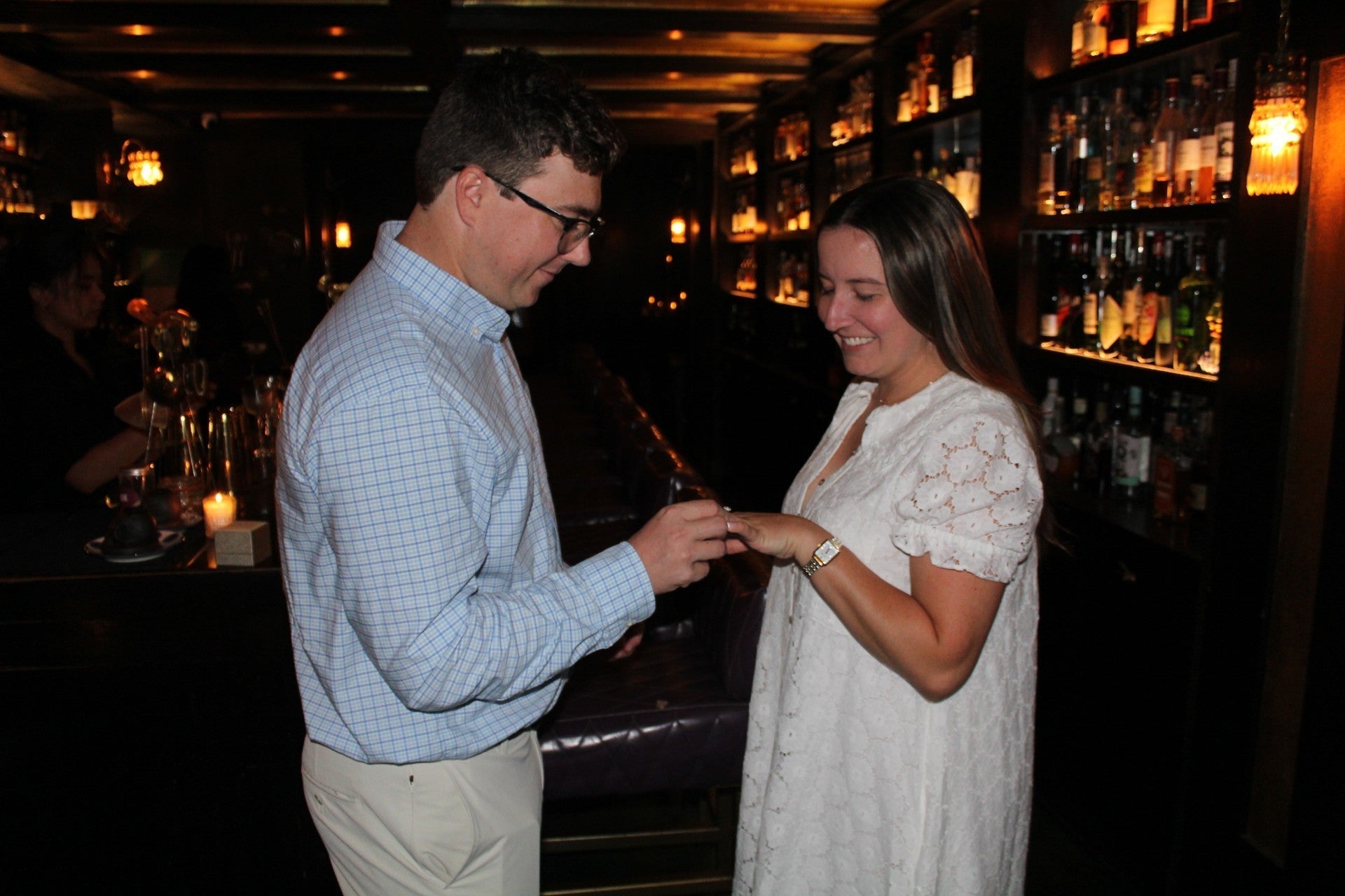 Man placing an engagement ring on a woman’s left hand in a dimly lit bar with backlit liquor shelves
