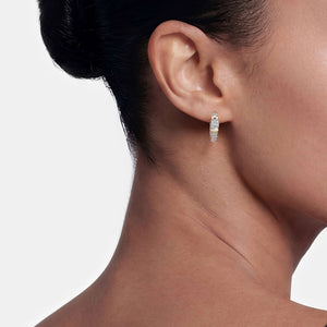 A close-up side profile of a woman's ear wearing a small silver huggie hoop earring with clear gemstones, set against a clean light gray background.