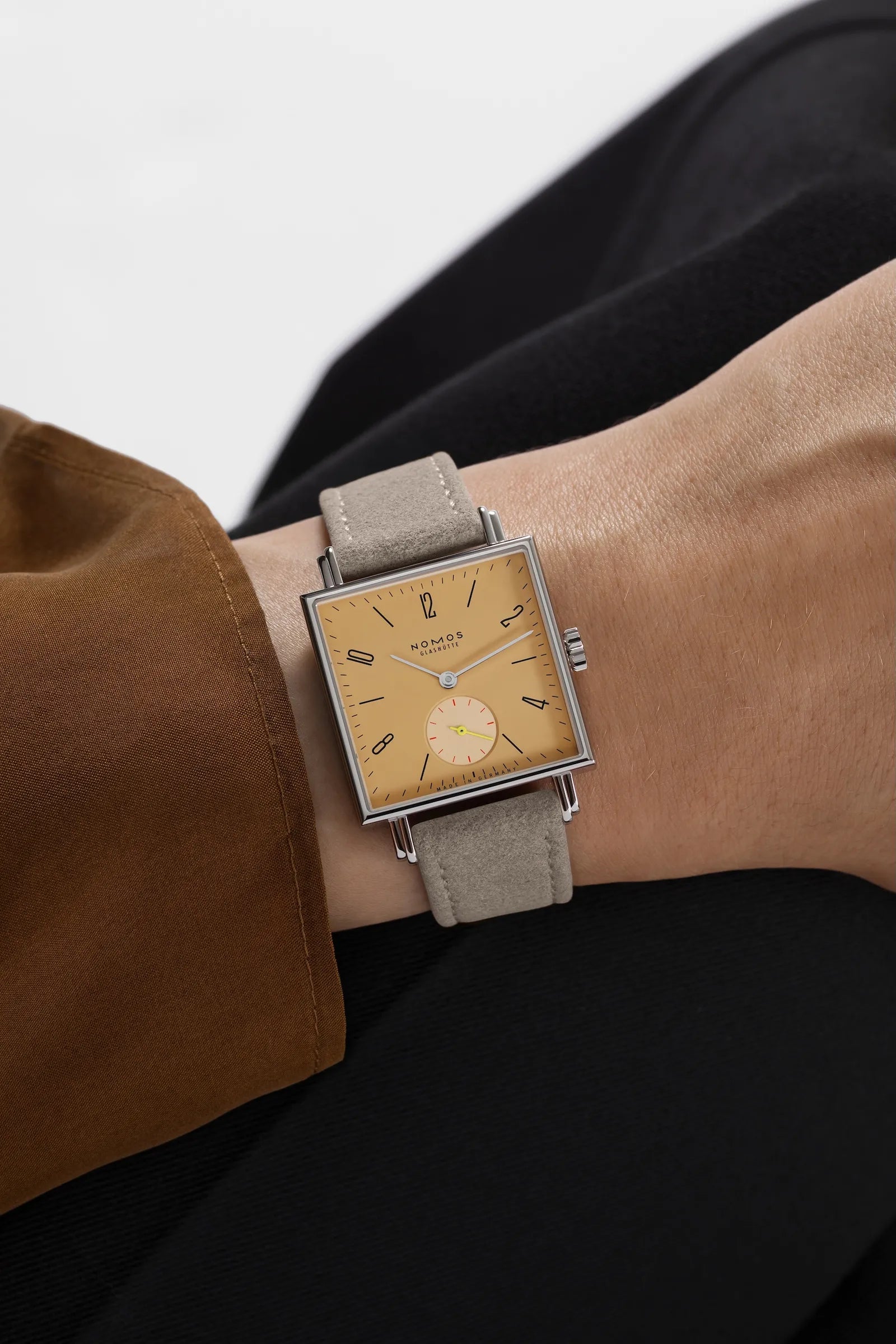 A wristwatch with a square silver-toned case, beige dial, and light taupe leather strap, shown worn on a person's wrist with a brown sleeve and black clothing visible.