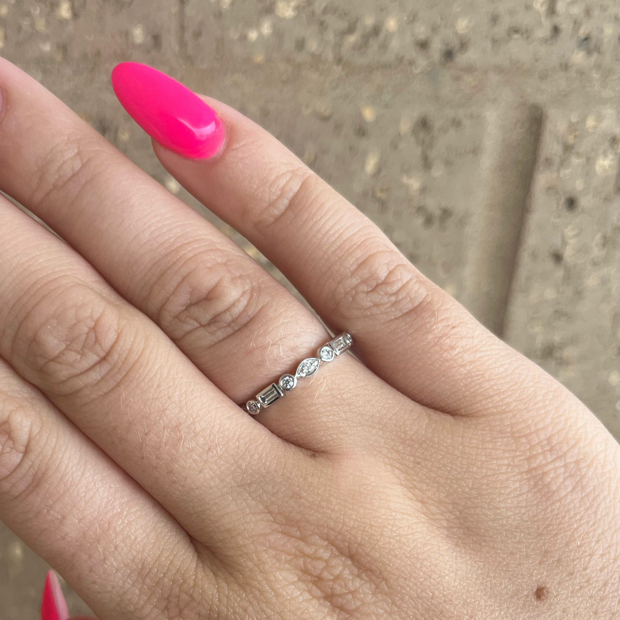 Delicate thin silver band with mixed-cut diamonds worn on a hand's ring finger; bright pink stiletto nail visible.