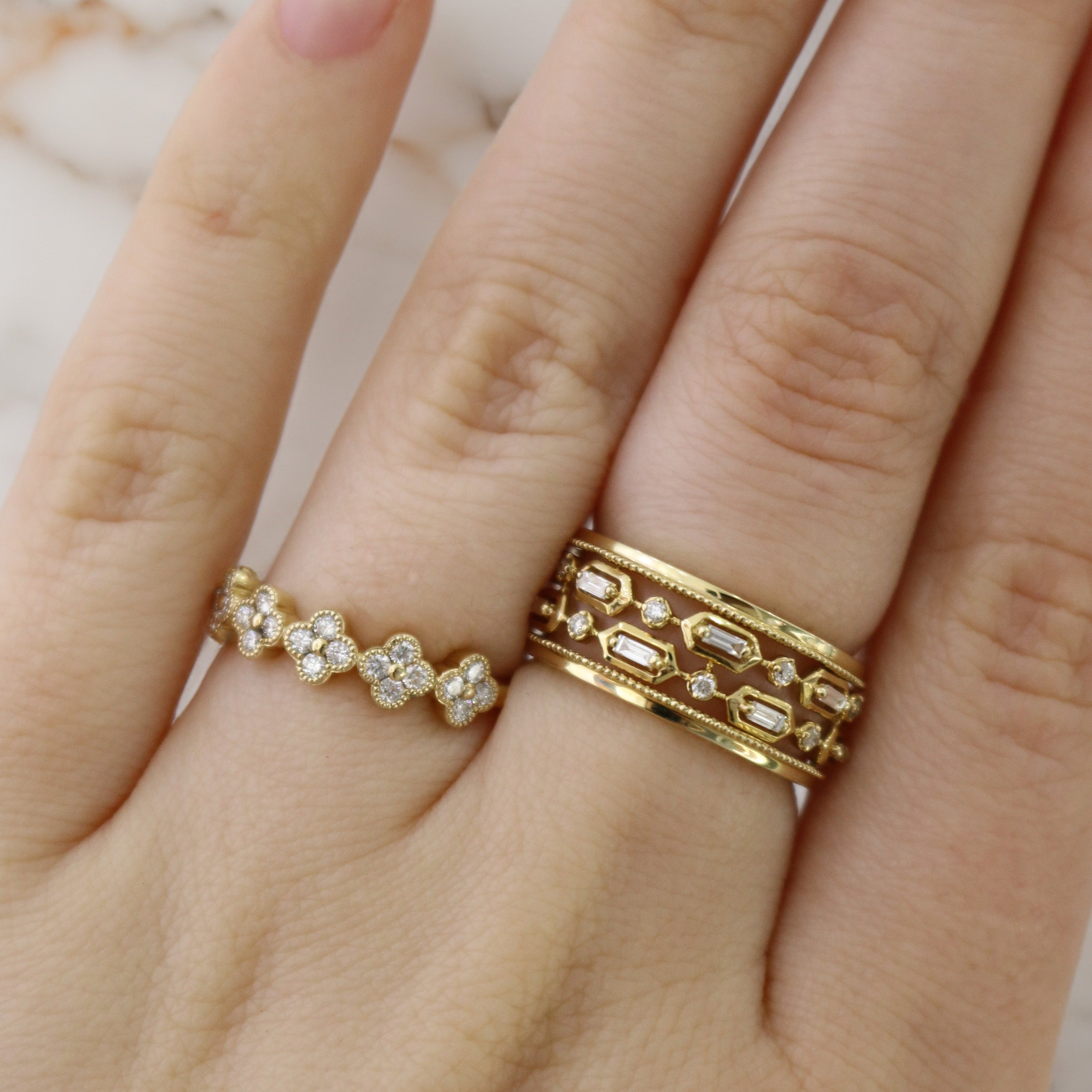 Two gold rings on a hand: a thin floral diamond band on the ring finger and a wide geometric diamond band on the middle finger