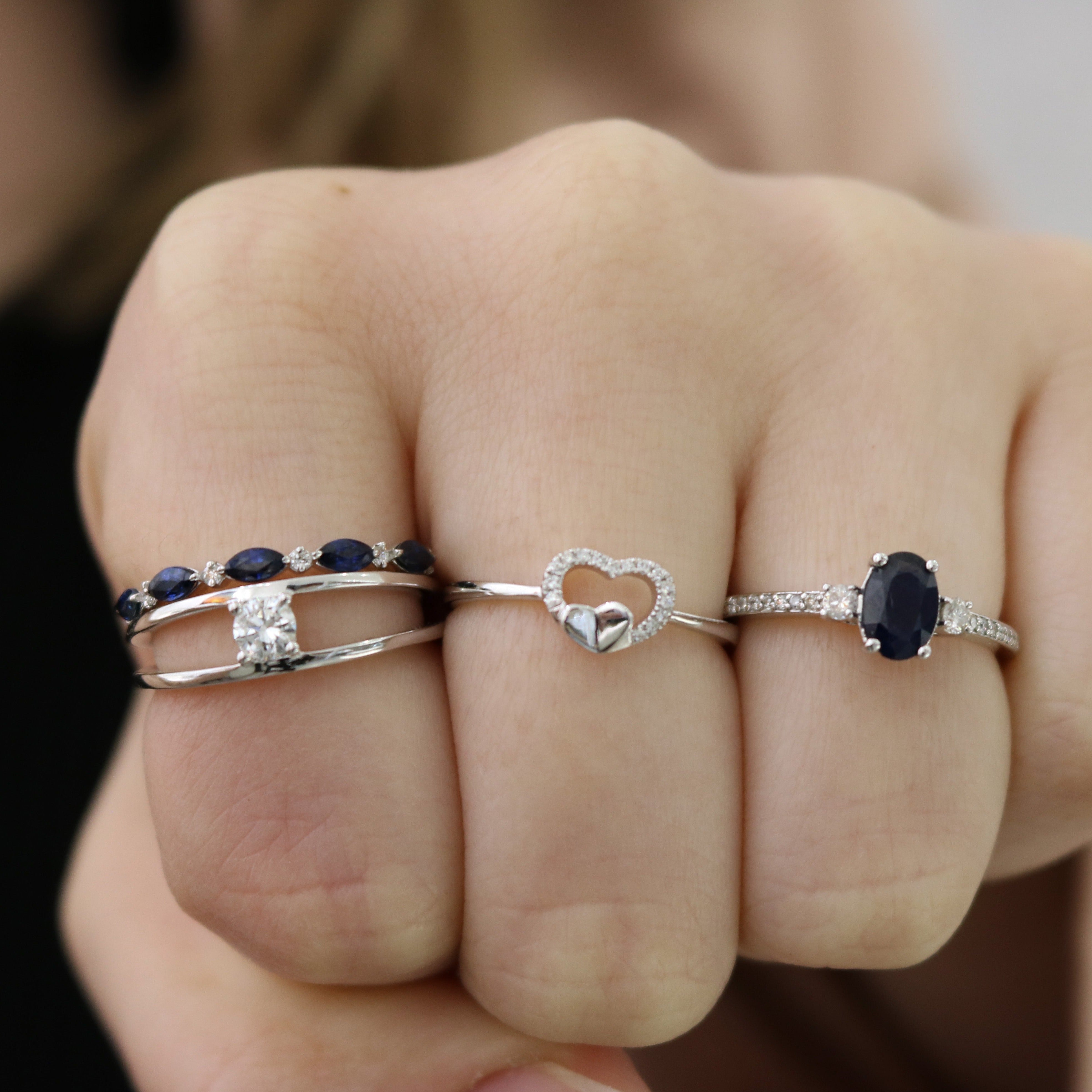Three silver rings on a hand: marquise blue-stone band, heart-shaped diamond-accent ring, and oval dark sapphire ring.