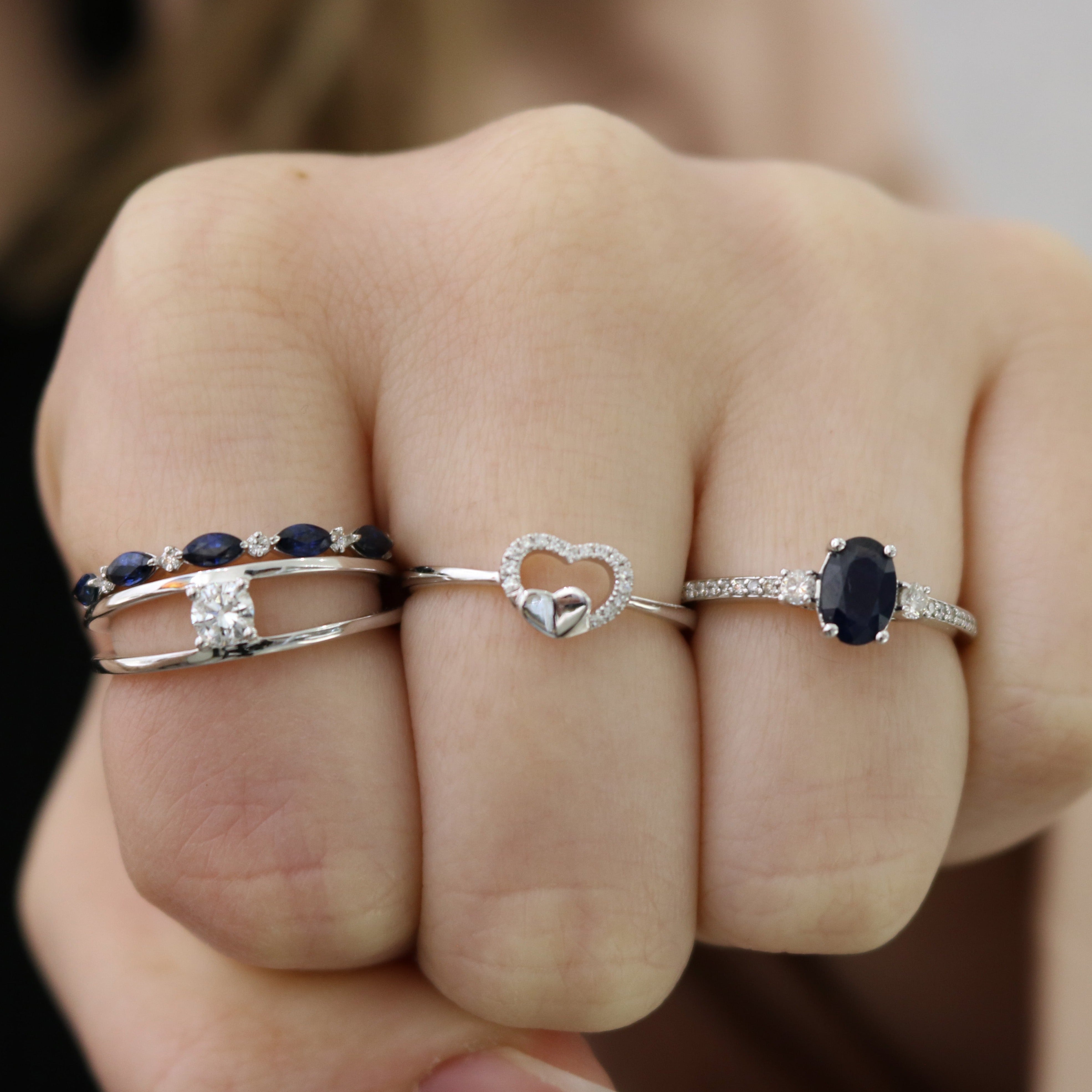 Hand wearing three silver rings: a band with marquise sapphires and diamonds, a small heart ring, and an oval sapphire ring.