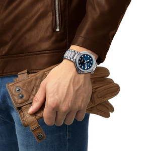 Person wearing brown leather jacket and jeans holding brown leather gloves wearing a Tissot watch with blue dial on a white background