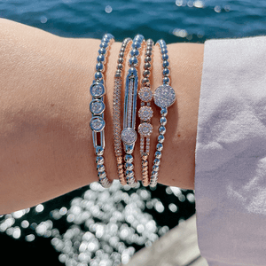 Stacked rose-gold and silver beaded bracelets with round diamond clusters, photographed on a wrist against sunlit water