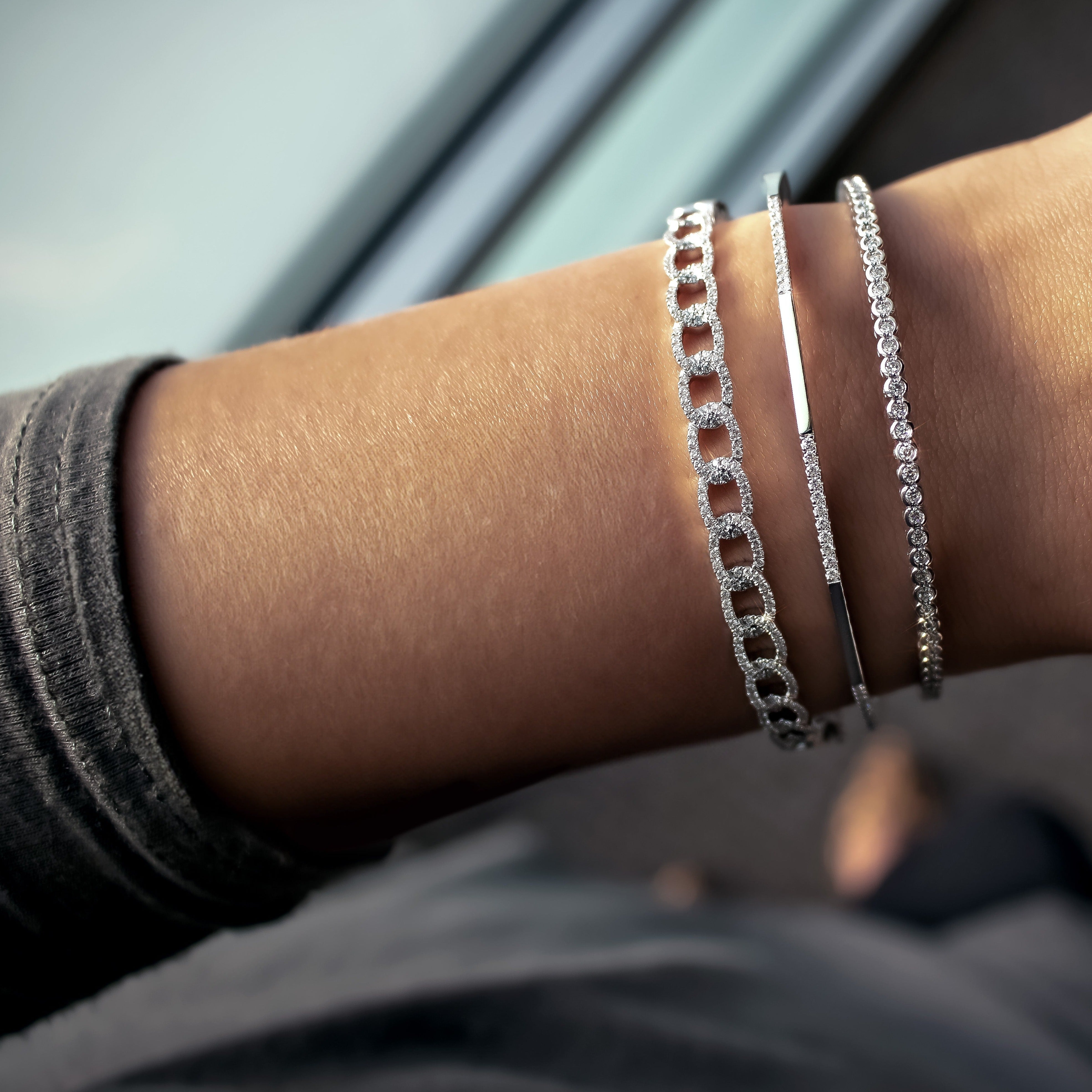 Three silver bracelets (chain, slim bar, and tennis styles) on a wrist beside a window.