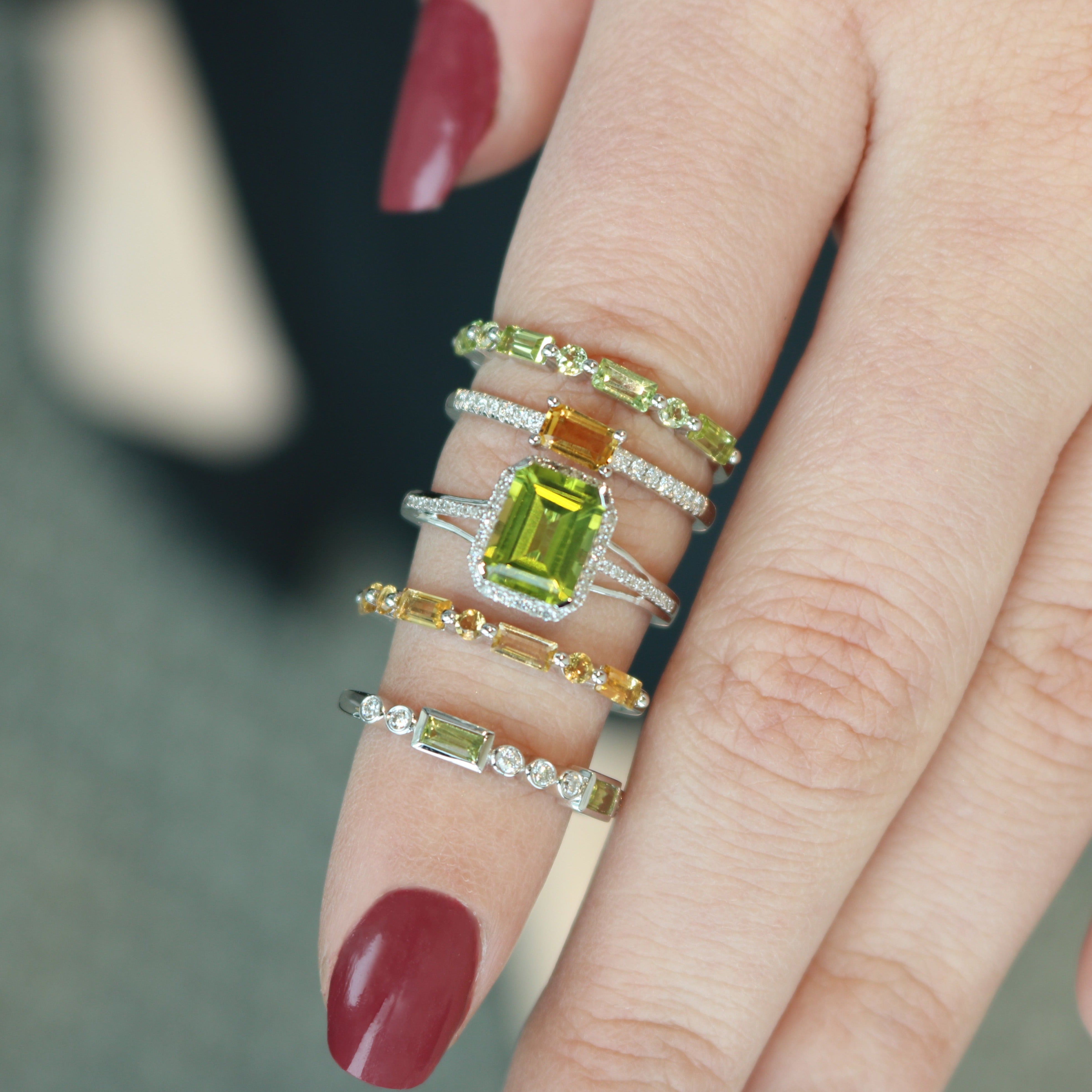 Finger wearing five stacked rings, center a rectangular green gemstone with smaller yellow-green stones; burgundy nail.