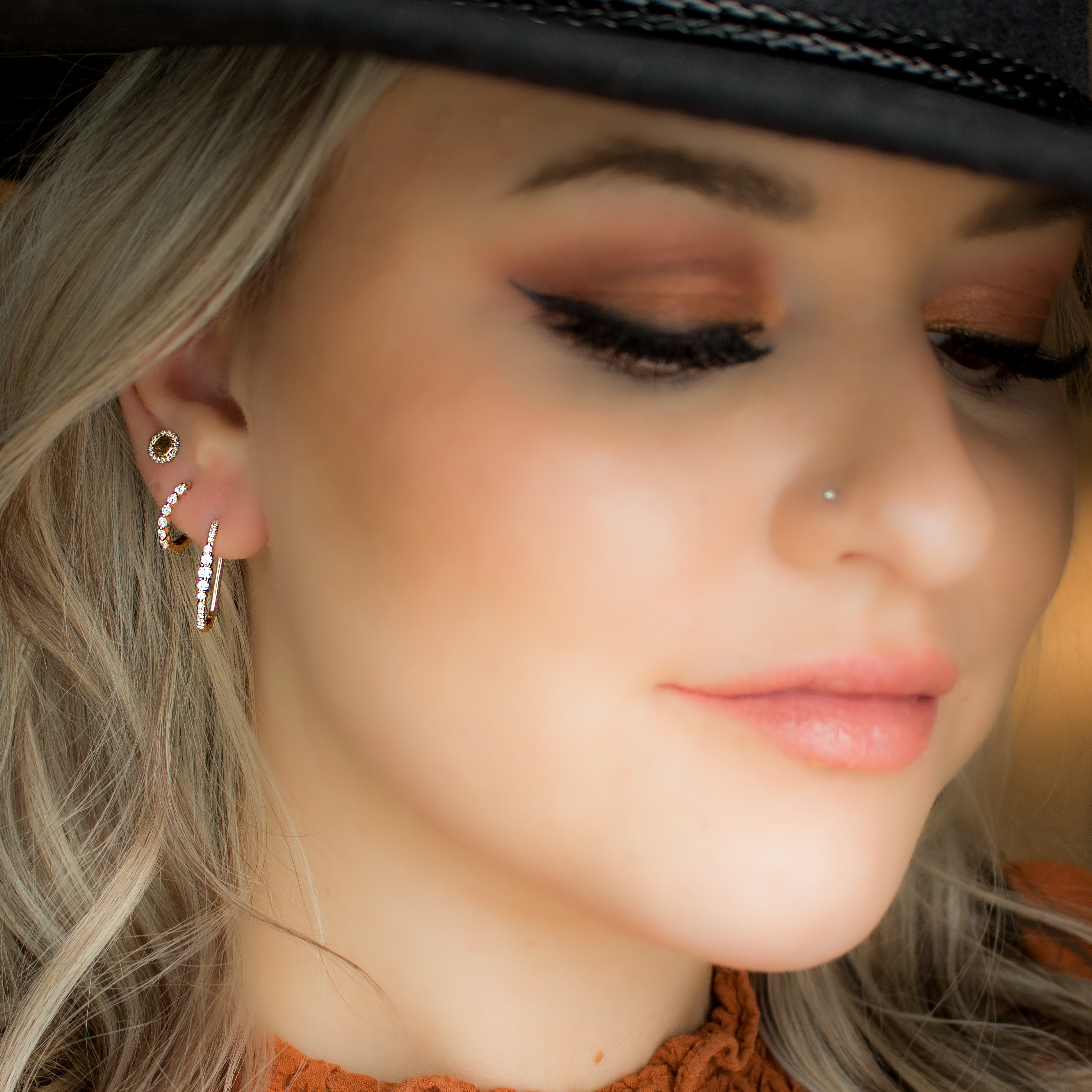 Close-up of woman's left ear wearing three gold diamond earrings (lobe and helix) and a small nose stud, with hat brim visible.