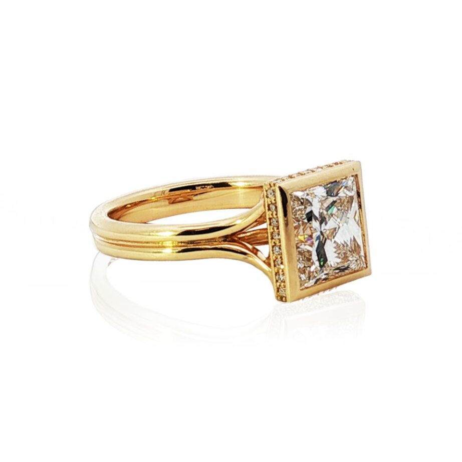 Side view of princess-cut diamond ring with square bezel-set center and pavé 18K yellow-gold band on white background