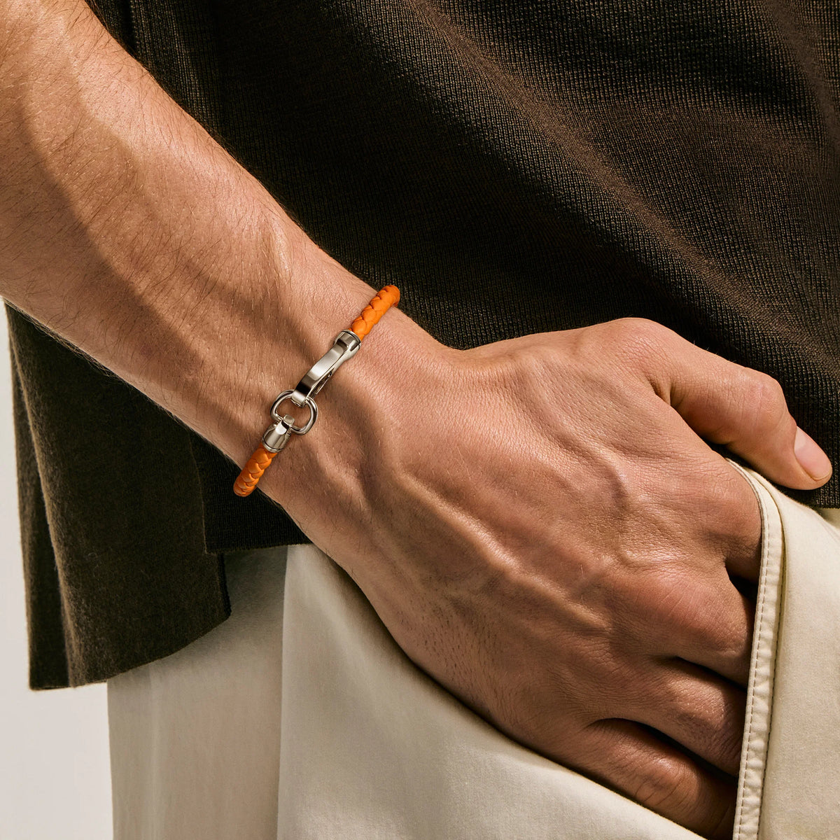 A close-up of a man's wrist wearing a bright orange beaded bracelet with a silver-toned clasp, set against a dark brown shirt and light beige trousers.