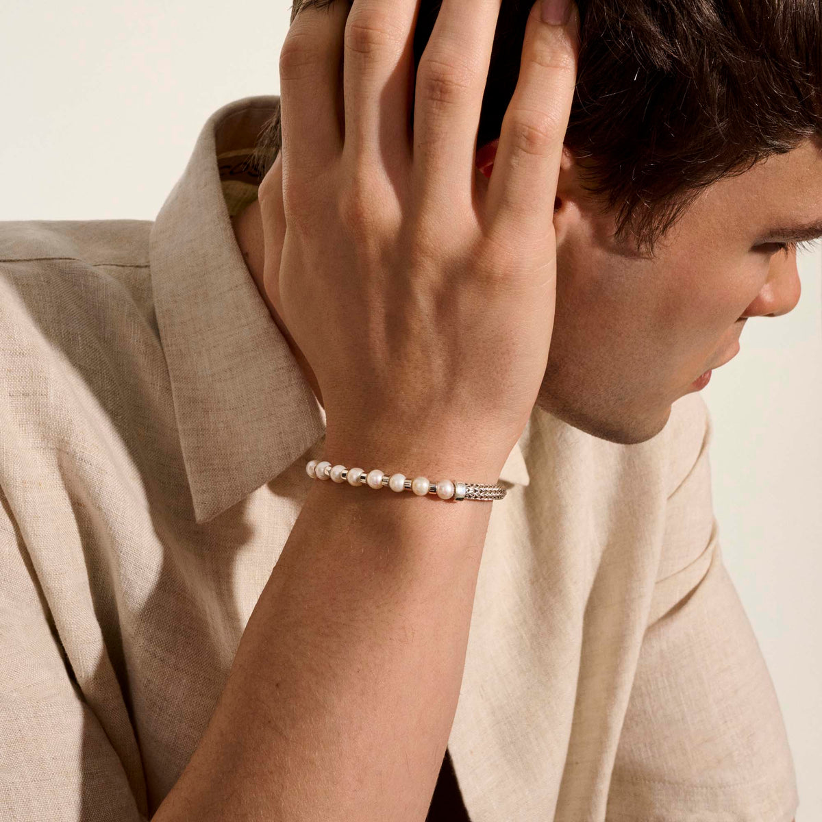 a man wearing a pearl and silver bracelet with his hand on his head wearing a beige shirt.