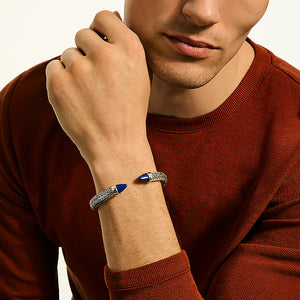 A close-up of a man wearing a silver-toned bangle bracelet with blue stone accents, paired with a rust-red knit sweater against a light background.