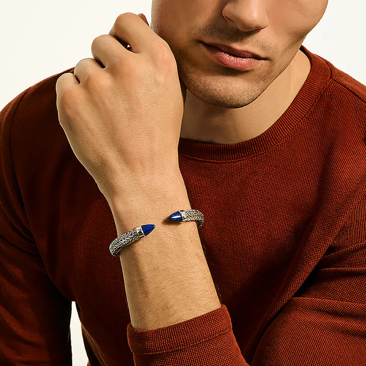 A close-up of a man wearing a silver-toned bangle bracelet with blue stone accents, paired with a rust-red knit sweater against a light background.