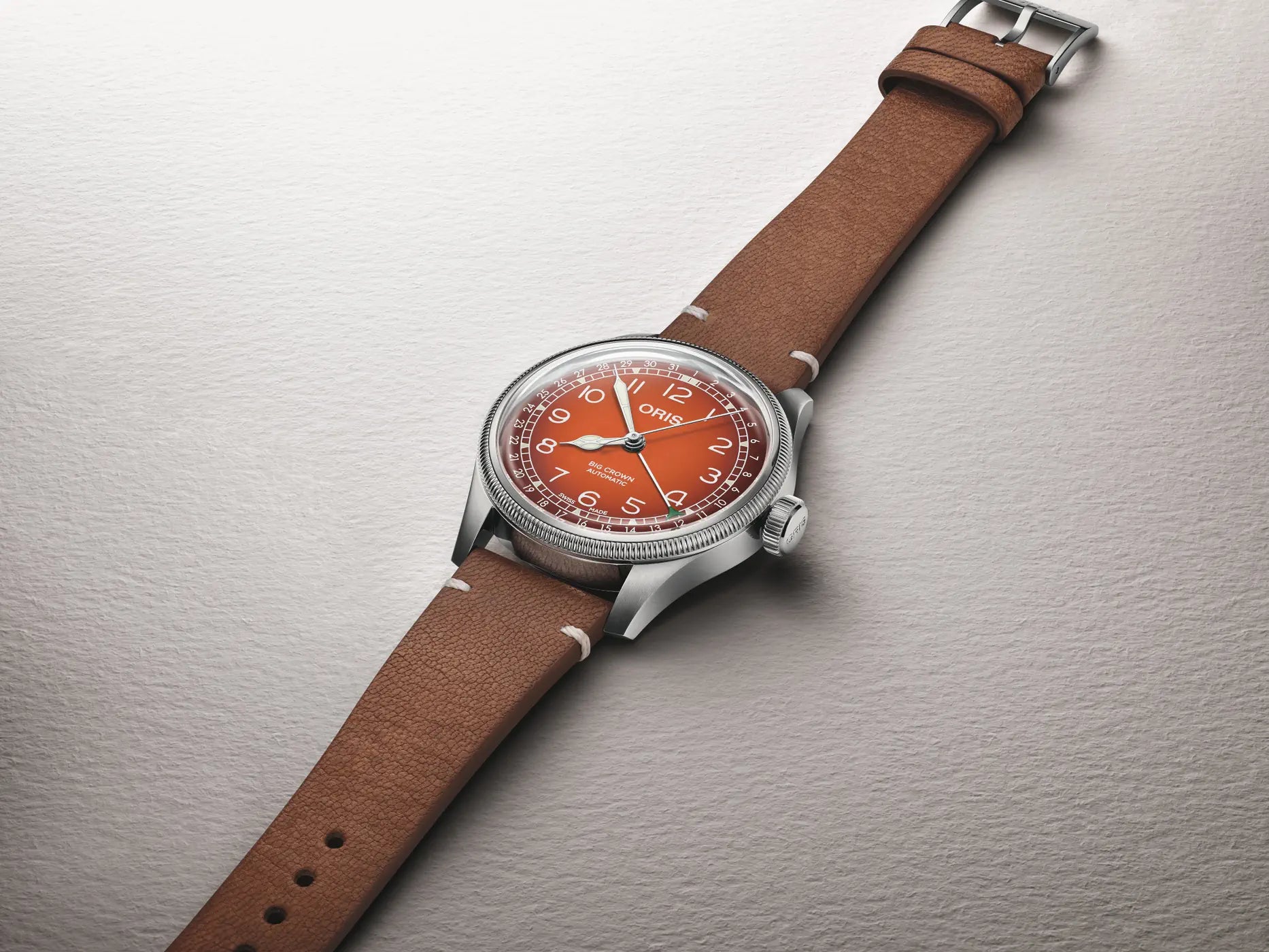 A wristwatch with a brown leather strap, silver-toned metal case, and a distinctive orange dial, photographed diagonally on a textured light gray background.