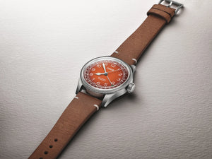 A wristwatch with a brown leather strap, silver-toned metal case, and a distinctive orange dial, photographed diagonally on a textured light gray background.