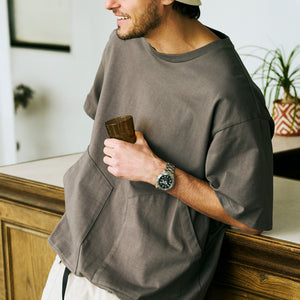 man standing next to counter wearing a olive green tshirt and holding a glass with a watch on his wrist