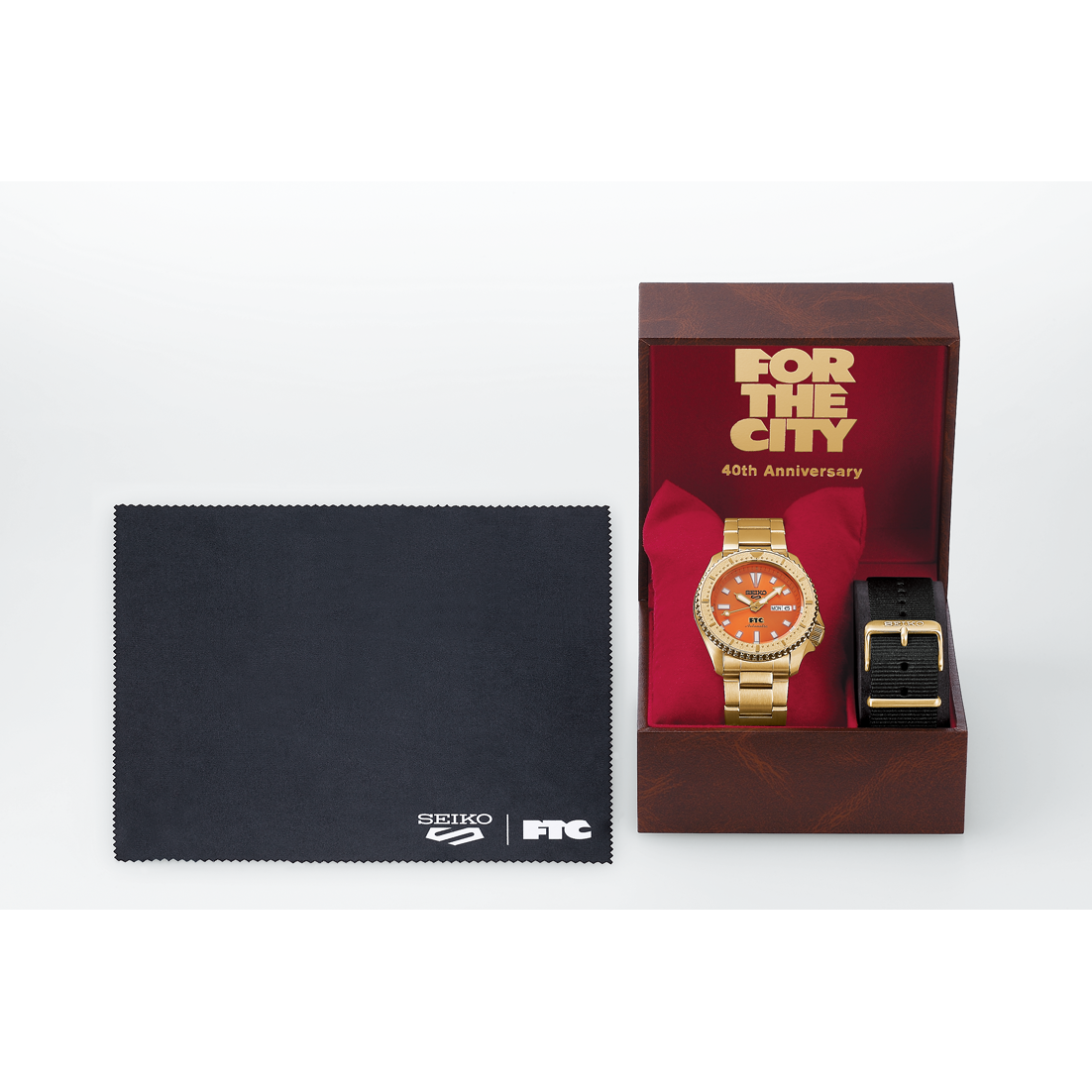 Gold watch with orange face and black strap in a box, accompanied by a Seiko cloth on a white background.