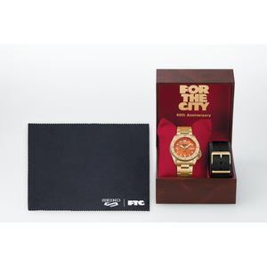 Gold watch with orange face and black strap in a box, accompanied by a Seiko cloth on a white background.