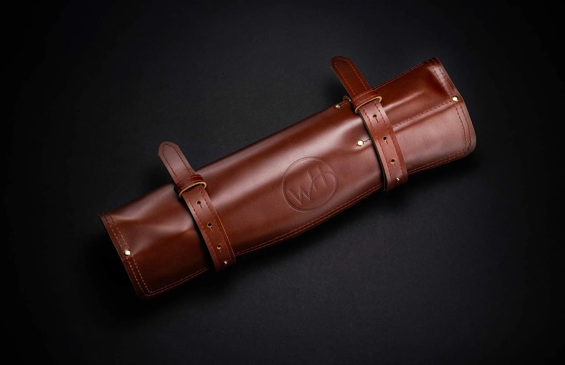 Angled top view of closed brown leather knife roll with embossed logo, two buckle straps and brass rivets on dark background
