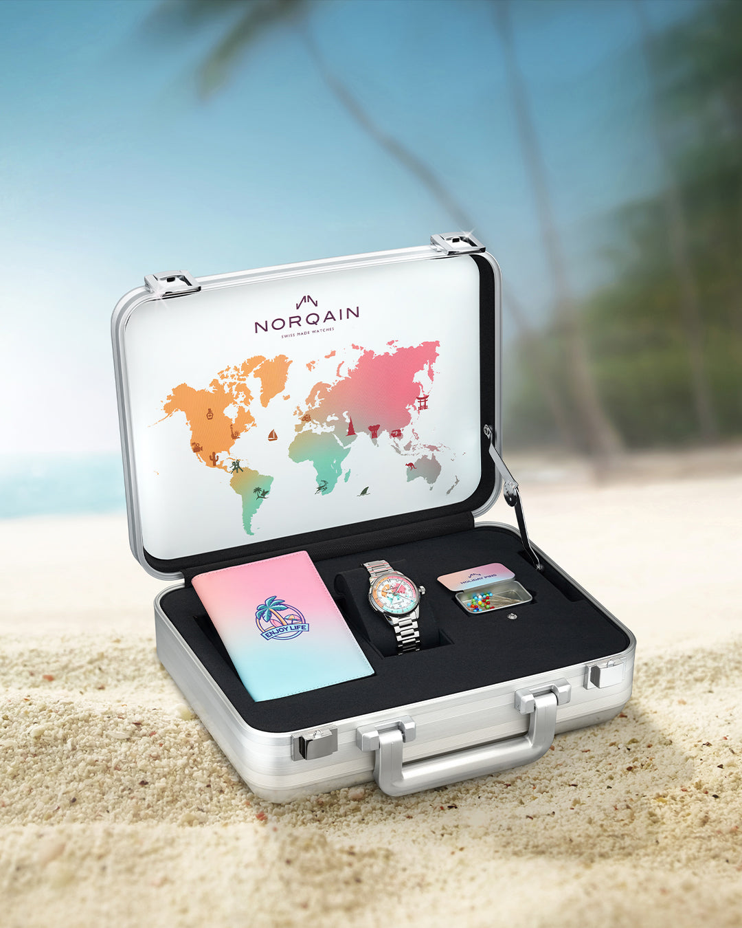steel case with watch and booklet sitting in case on sand with palm trees in the background with Norqain  written on the base