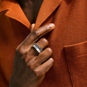 A close-up of a person's hand wearing a silver band ring with black lettering, resting on the lapel of an orange textured blazer.