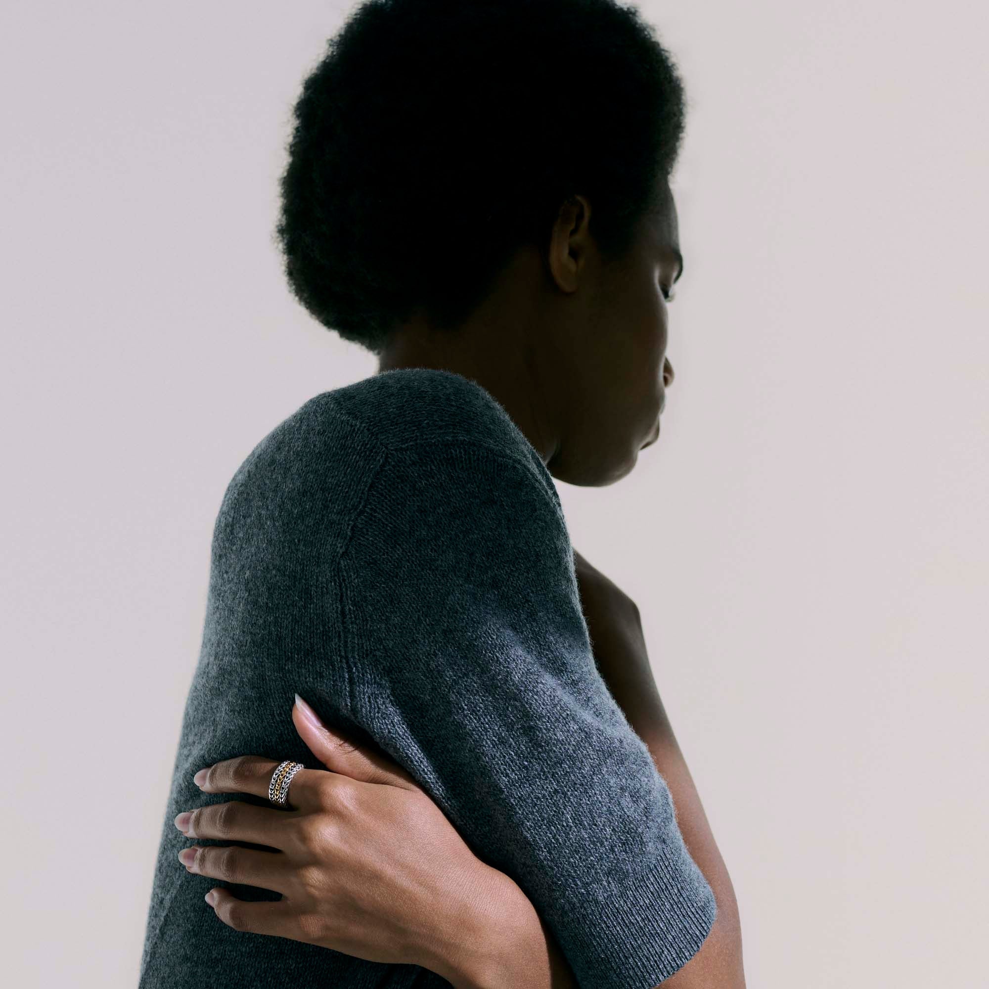 Person in gray short-sleeve sweater hugging herself; left hand rests on upper arm displaying a chain-link ring.