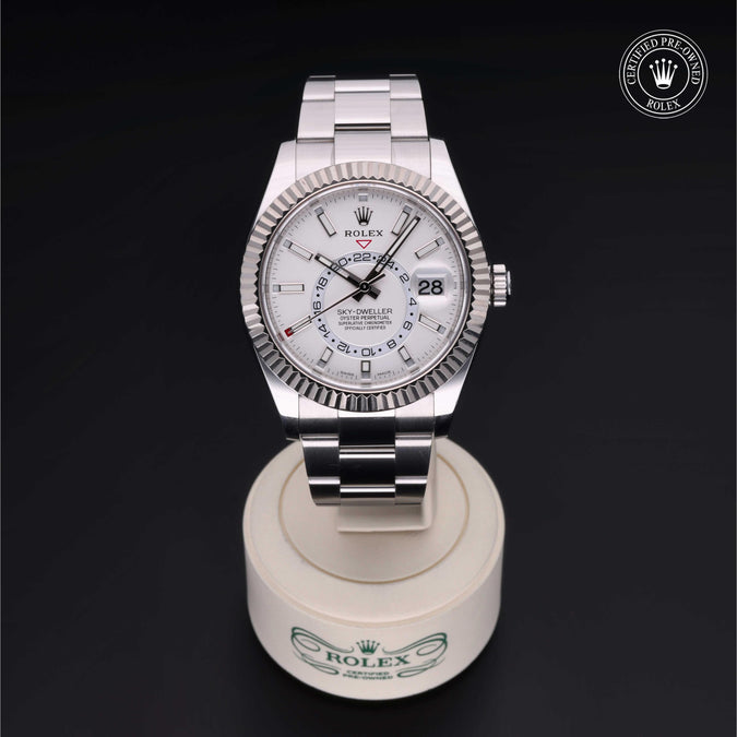 Front view of Rolex Sky-Dweller 42mm with white dial, fluted bezel and Oyster bracelet on white stand against dark background