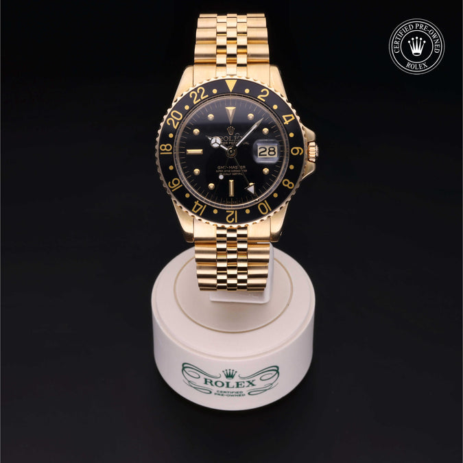 A gold-tone Rolex GMT-master watch with a black dial and black bezel featuring gold markings, sitting on a stand shown against a black background with a 'Certified Pre-Owned Rolex' emblem in the top right corner