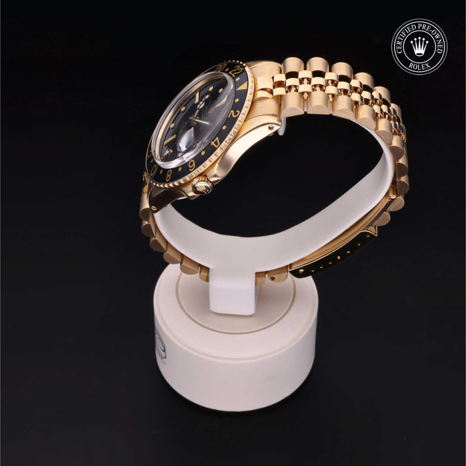 A gold-tone Rolex watch with a black dial and black bezel is displayed on a white stand against a black background. The Rolex logo is visible in the top right corner.