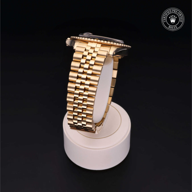 A gold-tone metal watch band displayed on a white cylindrical stand against a black background, with a Rolex crown logo watermark in the top right corner.