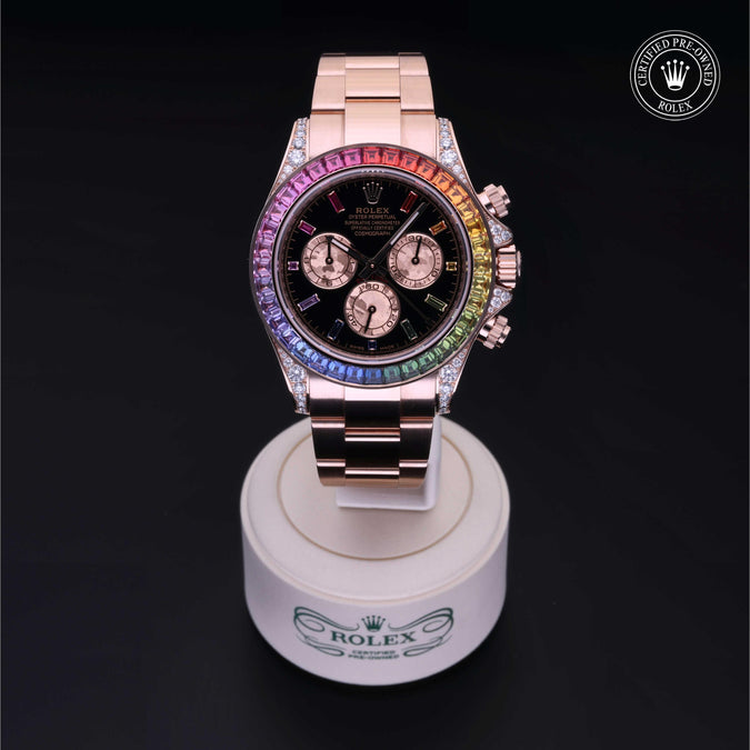 A luxury Rolex Daytona watch with a rose gold-tone case and bracelet, featuring a black dial and a vibrant rainbow-colored bezel. The watch is presented on a white stand and a solid black background with a 'Certified Pre-Owned Rolex' emblem in the top right corner.