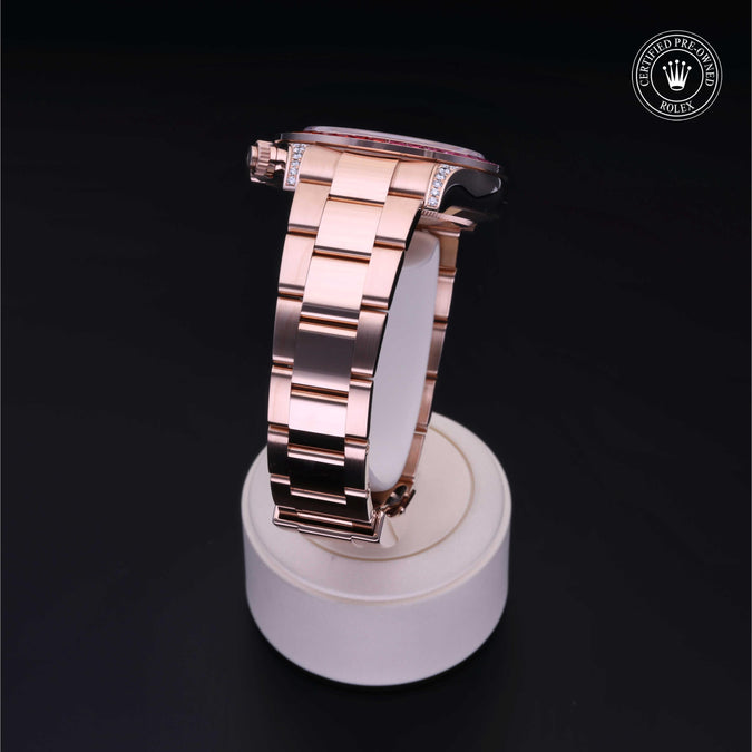 A rose gold metal watch with a matching link bracelet, displayed on a white stand against a black background. A Rolex crown logo watermark is visible in the top right corner.