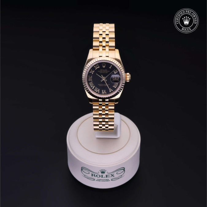 A gold-tone Rolex watch with a black dial and date window, displayed on a white Rolex-branded stand against a black background.