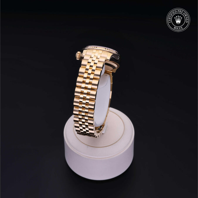 A gold-tone wristwatch with a matching metal link bracelet displayed upright on a round white stand against a black background. A Rolex crown logo watermark appears in the top right corner.