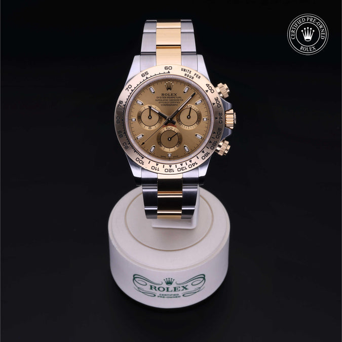 Rolex Daytona watch with a gold dial and two-tone steel and gold Oyster bracelet, displayed on a white Rolex-branded stand against a black background.