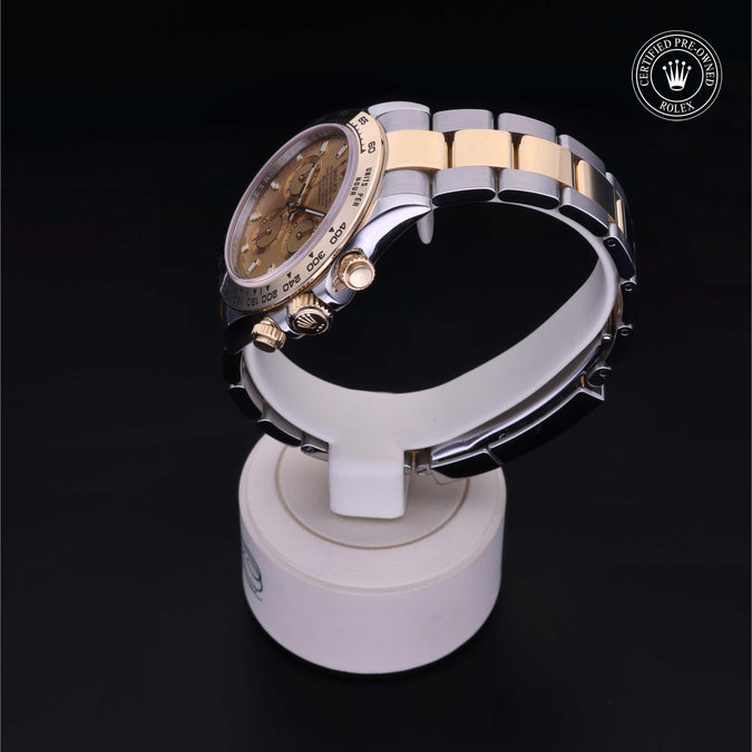A two-tone Rolex Daytona watch with a gold dial and integrated bracelet, displayed on a white stand against a black background. A Rolex logo watermark is visible in the top right corner.