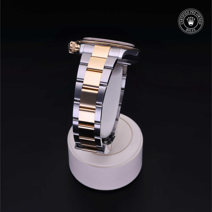 A two-tone stainless steel and gold watch band displayed on a white cylindrical stand against a black background, with a Rolex crown logo watermark in the top right corner.