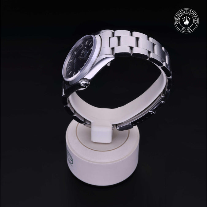 A stainless steel watch with a dark dial and silver link bracelet, displayed on a white cylindrical stand against a black background. A circular Rolex crown logo watermark is visible in the top right corner.