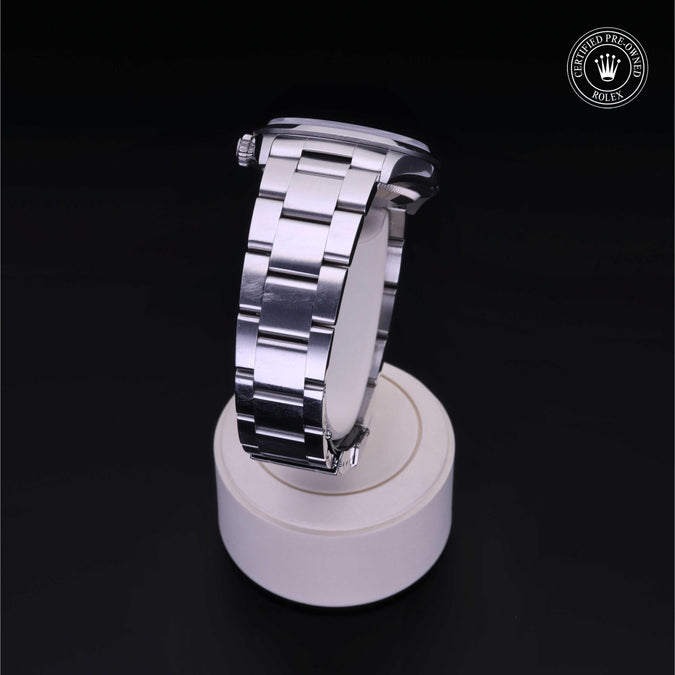 A stainless steel watch with silver link bracelet, displayed on a white cylindrical stand against a black background. A circular Rolex crown logo watermark is visible in the top right corner.