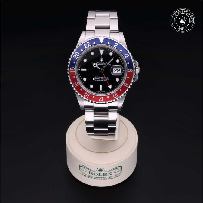 A Rolex GMT-Master II watch with a silver Oyster bracelet and a black dial featuring a red and blue two-tone bezel, displayed on a white Rolex-branded stand against a black background.