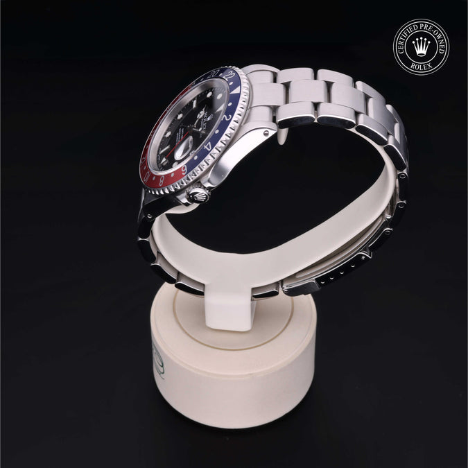A Rolex GMT-Master II watch with a silver Oyster bracelet and a black dial featuring a red and blue two-tone bezel, displayed on a white Rolex-branded stand against a black background.