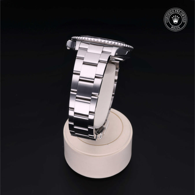 A silver stainless steel watch band with a polished metal link design, shown resting on a round beige display stand against a black background. A circular Rolex logo watermark is visible in the top right corner.