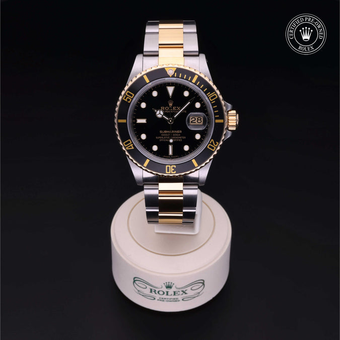 A Rolex Submariner watch with a two-tone steel and gold bracelet, featuring a black dial and a black and gold rotating bezel, sitting on a white stand set against a solid black background with a 'Certified Pre-Owned Rolex' emblem in the top right corner.