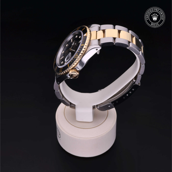 A two-tone Rolex watch with a gold and silver metal bracelet, black dial, and gold bezel, displayed on a white stand against a black background. The Rolex logo is visible in the top right corner.