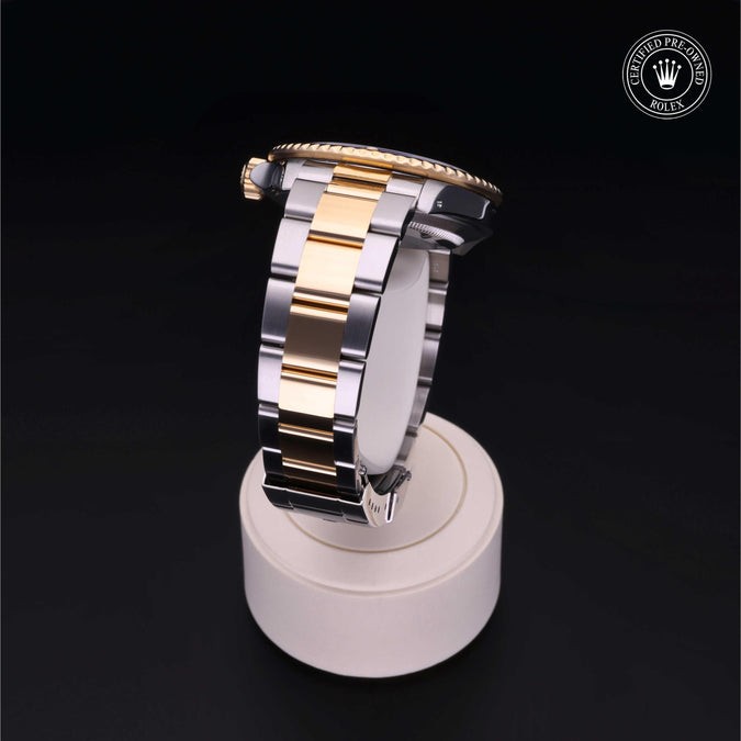 A two-tone stainless steel and gold watch band displayed on a white circular stand against a black background, with a Rolex crown logo watermark in the top right corner.