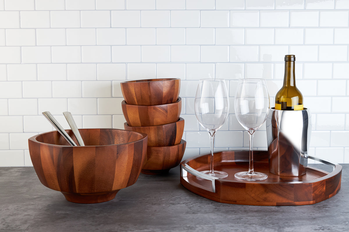 Front view of Rivet acacia wood salad bowl with stainless steel servers, stacked matching bowls and tray on gray countertop