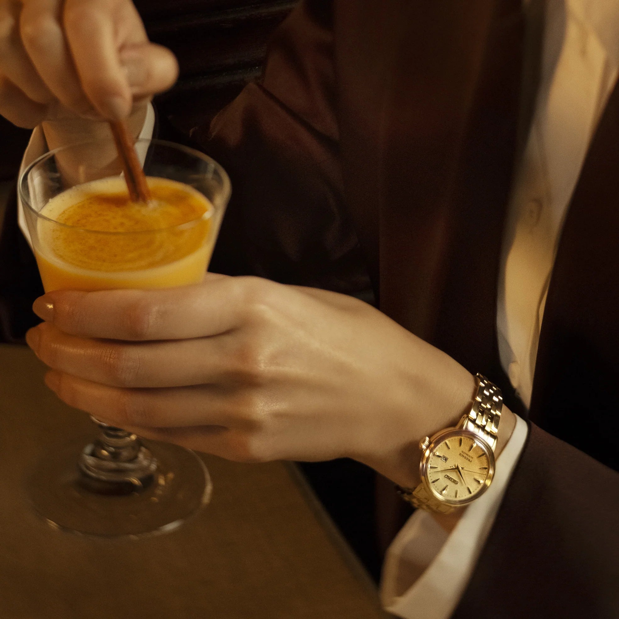 Lady holding a warm cocktail wearing a gold watch with a brown jacket