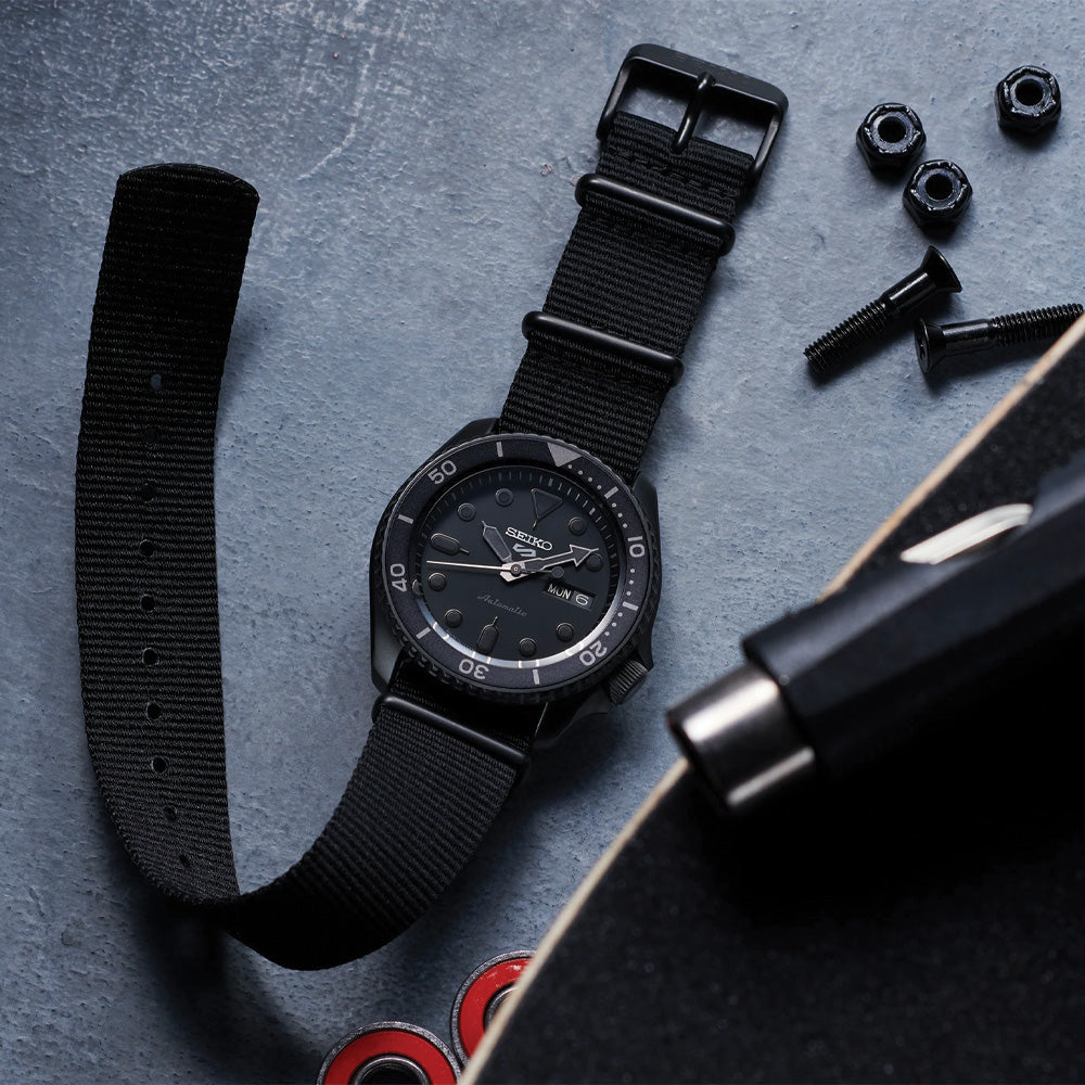 Flat lay of Seiko SRPD79 watch with black ion-coated case, black nylon strap, rotating bezel and day/date on gray background